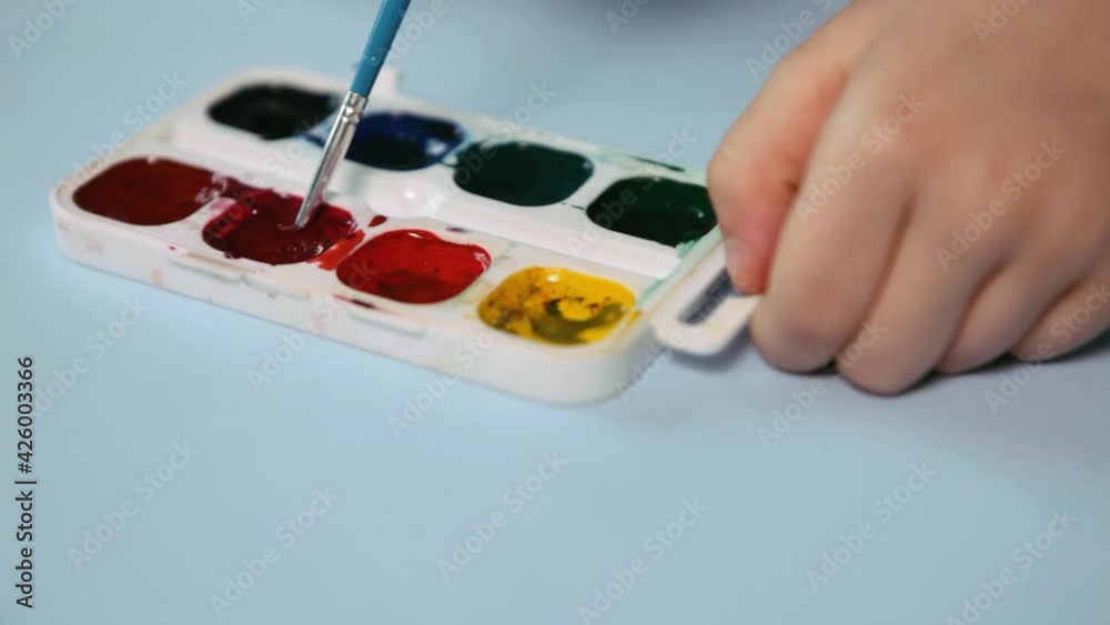 Child painting Closeup. The palette of colors is on the blue table. A child's hand holds a brush and mokes it in red High quality FullHD footage