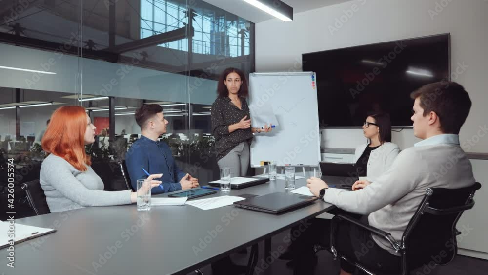 Black woman reporting on progress while standing by flipchart with hand-drawn growth graph at business meeting. Multiethnic team analyzing financial success and planning further strategy Black woman reporting on progress while standing by flipchart with hand-drawn growth graph at business meeting. Multiethnic team analyzing financial success and planning further strategy