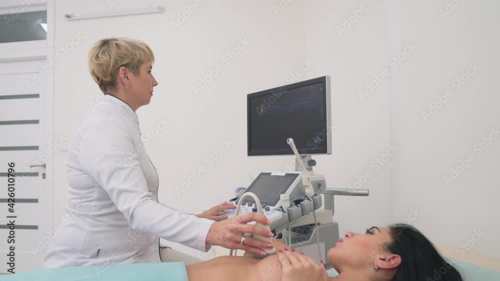 Side view of middle aged mammologist using ultrasound device to examine naked breast of female patient. Young woman lying on coach in clinic office and covering nipple with hand. Concept of medicine. Side view of middle aged mammologist using ultrasound device to examine naked breast of female patient. Young woman lying on coach in clinic office and covering nipple with hand. Concept of medicine.