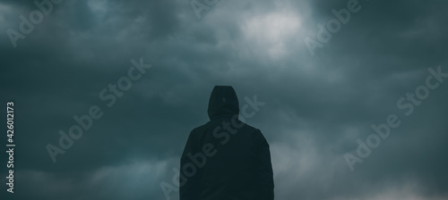 Rear view of male person wearing hooded jacket against dark moody dramatic clouds at sky photo