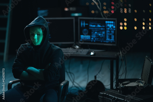 Dangerous Internationally Wanted Hacker with Masked Face Speaks into the Camera. In the Background His Operating Room with Multiple Displays and Cables.