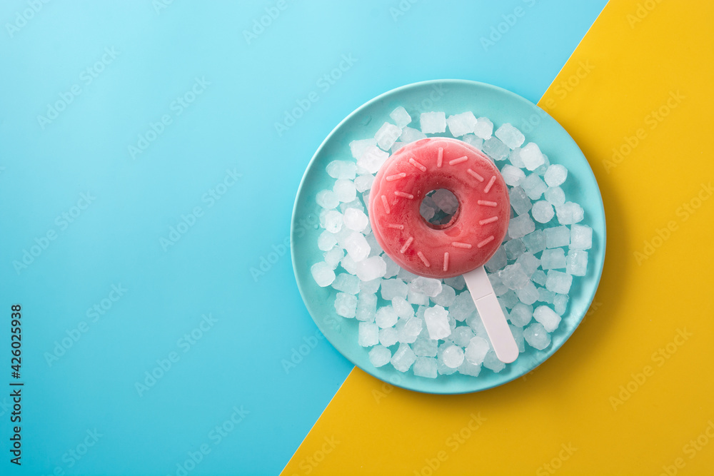 Obraz premium Strawberry donut popsicle and crushed ice on yellow and blue background. Copy space