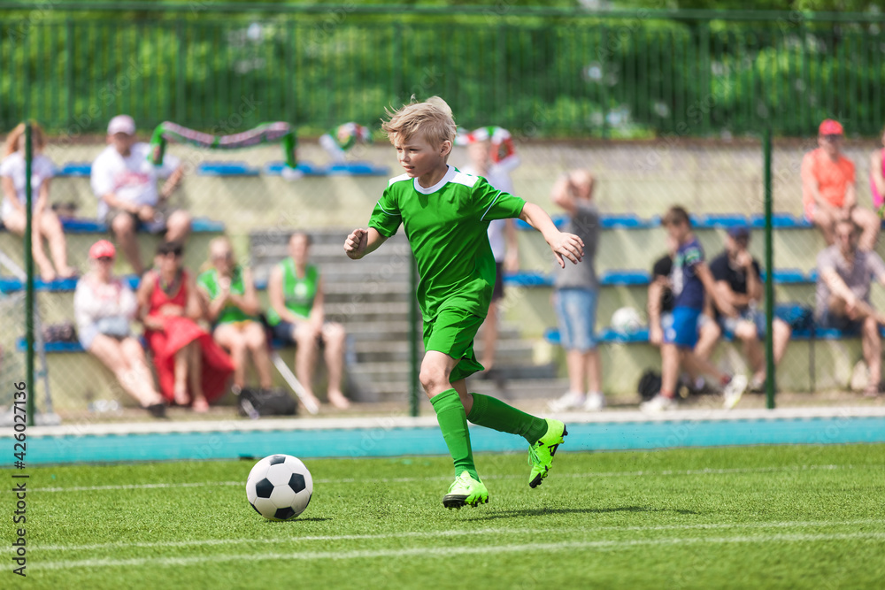 School Boy Running Football Ball on Stadium Field. Happy Boy Soccer Player Running Fast Ball on Tournament Competition Match. Kid in Green Sports Jersey Kit: Shirt, Shorts, Socks and Cleats