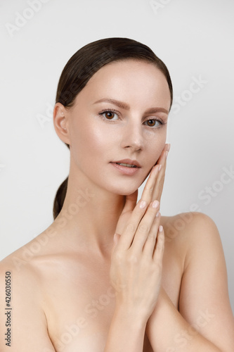 Young woman with beautiful healthy face - isolated on white background, studio hi-end shot, skin care concept. Hands and fingers gently touch facial skin, brown hair parted, gathered in low ponytail