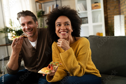 Cheerful young couple sitting on sofa at home. Happy woman and man eating pizza while watching a movie..