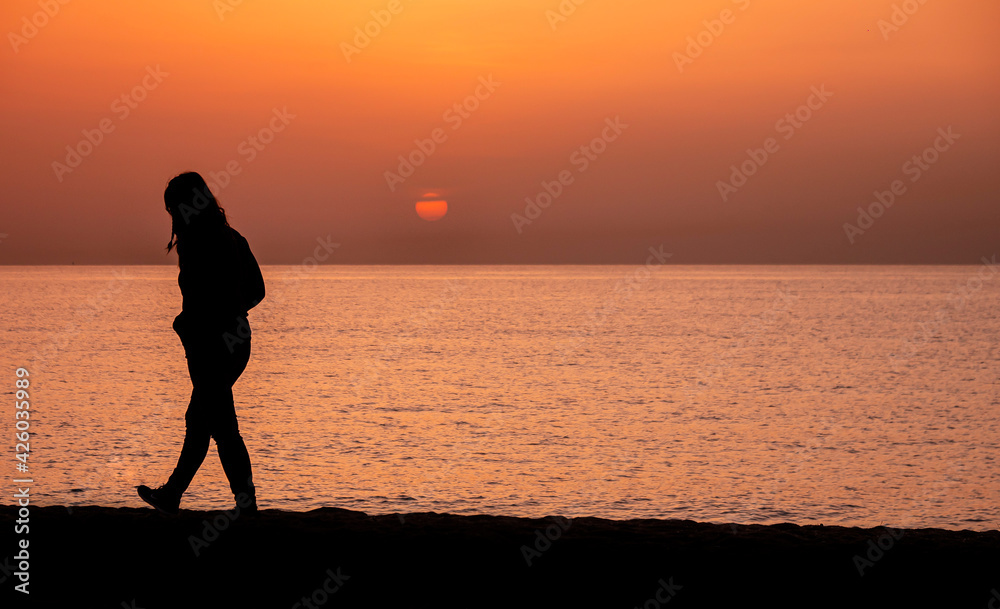 Fototapeta premium Silhouette of a woman walking by the ocean. Woman shilouette at sunrise on the beach