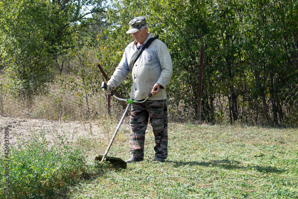 An old man mows grass with gasoline trimmer. Sunny autumn day. Front view. An old man mows grass with gasoline trimmer. Sunny autumn day. Front view.