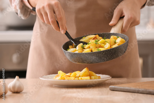 Woman putting tasty potatoes from frying pan into plate in kitchen