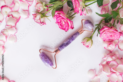 Fototapeta Naklejka Na Ścianę i Meble -  The front roller is made of amethyst on a white background with pink roses. Massager for lifting the skin made of natural stone. Beauty tools.