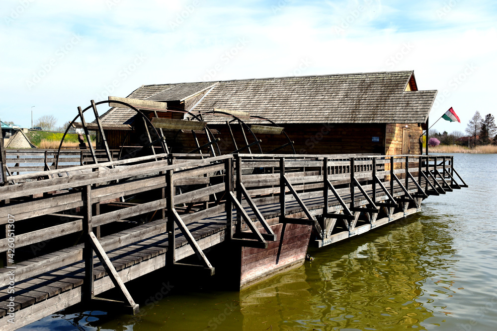 old, historic wooden floating boat mill on green river water. large wooden boat house like structure with huge spinning mill wheels and plank blades. blue sky above. scenic scene. reflections