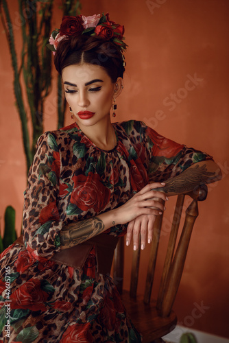 Portrait of beautiful dark-haired tattooed woman with crown braid hairstyle and floral head wreath wearing chic chiffon dress sitting on the wooden chair among cactuses. Mexican. Frida Kahlo image