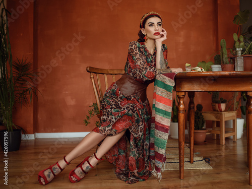 Portrait of beautiful dark-haired tattooed woman wearing precious headband, earrings and chic chiffon dress with basque belt sitting on the retro wooden chair at the table with many cacti around photo