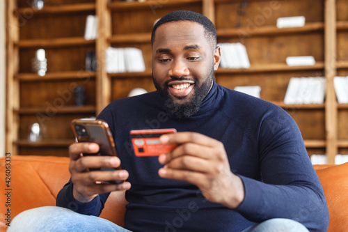 Smiling African-American man shopping online sitting on the sofa at home, holding credit card and smartphone, dark skinned multiracial guy making food order, purchasing online, booking and paying
