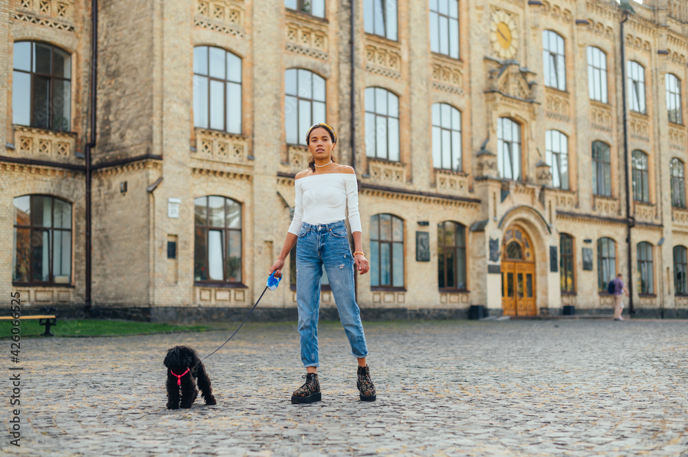 Beautiful hispanic woman walking with a little black dog on a leash on the street against the backdrop of a historic old building, looking at the camera.