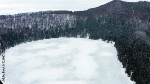 Volcanic Crater Lake Of Saint Anne Covered In Ice And Surrounded With Pine Forest At Winter In Harghita, Romania. - aerial photo