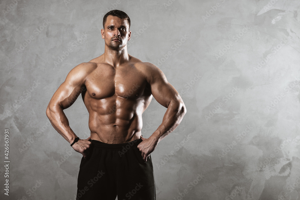 Fototapeta premium Fitness in gym, sport and healthy lifestyle concept. Handsome athletic man with naked torso demonstrates his muscles and trained body. Bodybuilder male model posing on grey background.