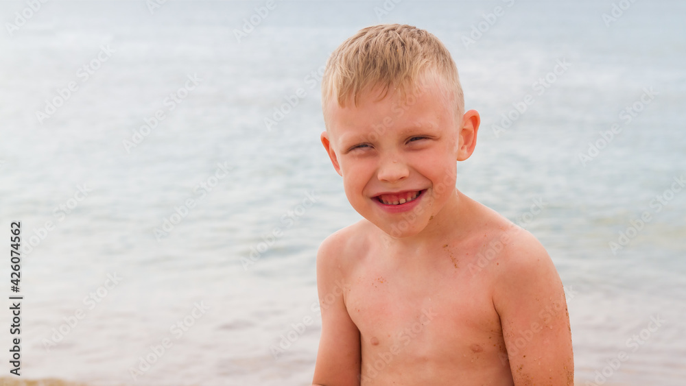 Funny caucasian boy sits on the beach by the sea in summer and laughs Funny caucasian boy sits on the beach by the sea in summer and laughs