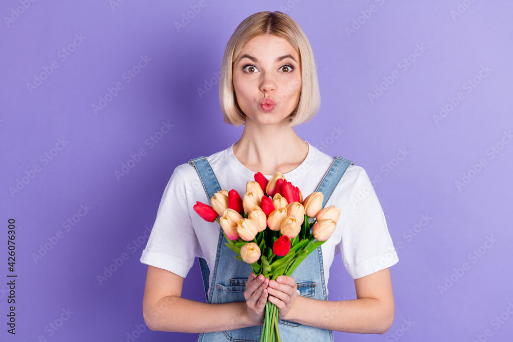 Photo of nice optimistic short hairdo blond lady hold flowers blow kiss wear white t-shirt isolated on violet background Photo of nice optimistic short hairdo blond lady hold flowers blow kiss wear white t-shirt isolated on violet background