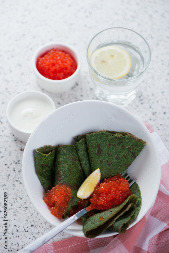 White bowl with spinach crepes, red caviar and sour cream, studio shot on a beige stone background, selective focus White bowl with spinach crepes, red caviar and sour cream, studio shot on a beige stone background, selective focus