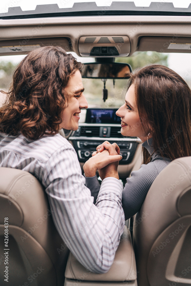 Happy young couple hand in hand in front of car, modern car interior, looking at each other and smiling Happy young couple hand in hand in front of car, modern car interior, looking at each other and smiling