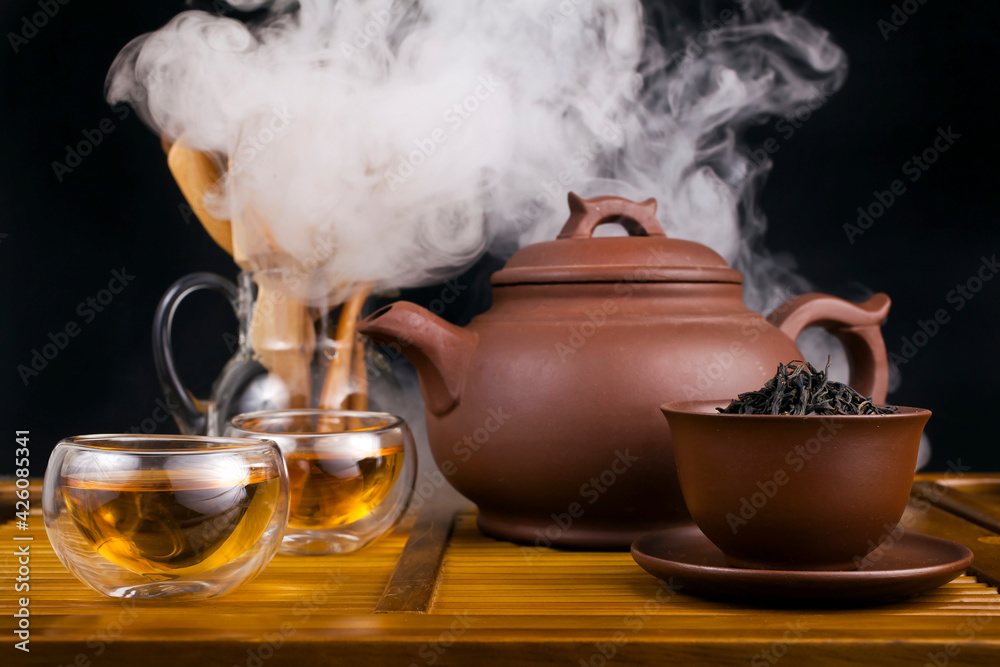 Chinese tea ceremony. Clay teapot with smoked red tea Lapsang souchong on a  black background, glass cups of hot tea with vapour. Stock Photo