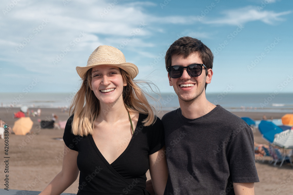 Young couple laughing and looking at the camera with the beach in the background.