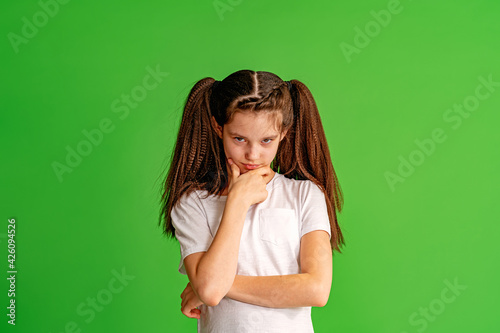 little girl looks serious from under brows, hands folded and brows furrowed. child unhappy and offended, shows disapproval and holds her chin in her hand against green background