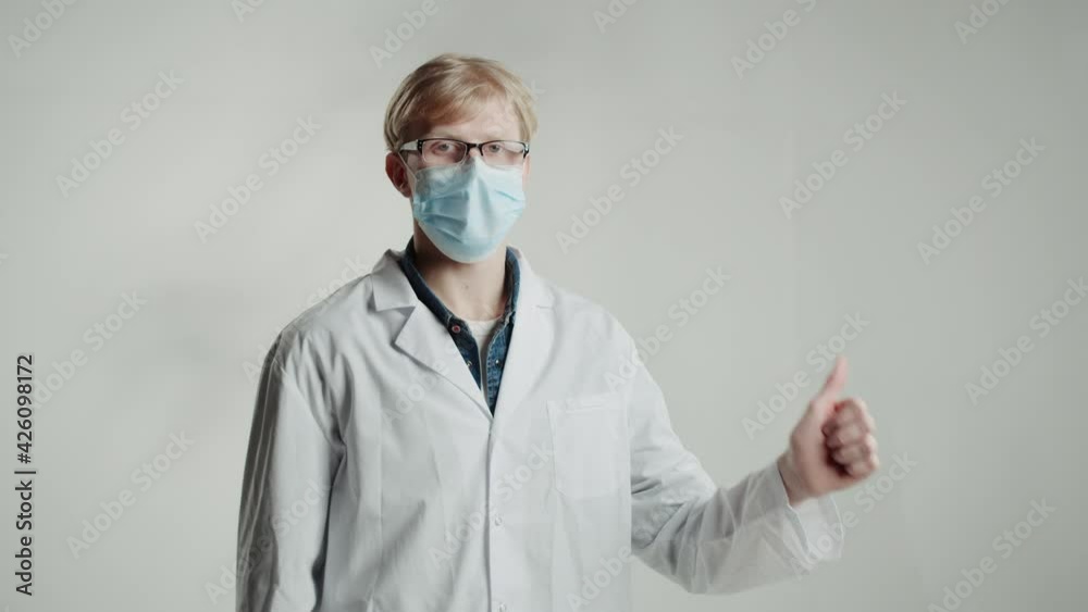 Caucasian man doctor in medical mask and glasses, showing thumbs up, gesturing, looking in camera on white backgrounds. Student of medical university portrait, young blonde nurse male.