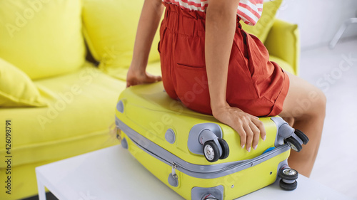 partial view of woman sitting on yellow travel bag near sofa in living room