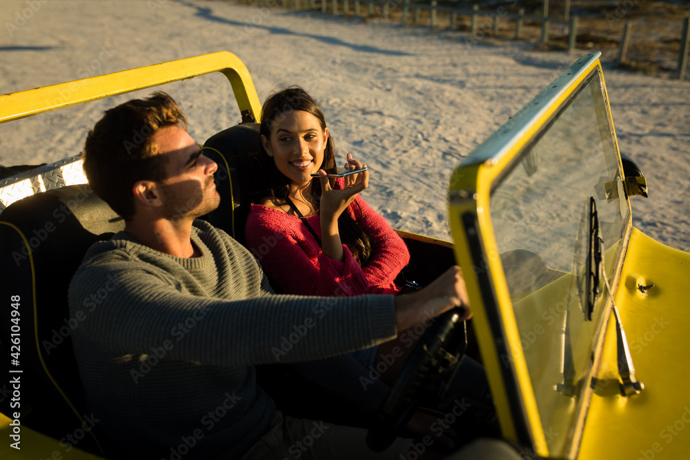 Caucasian couple sitting in beach buggy by the sea during sunset talking by phone - obrazy, fototapety, plakaty 