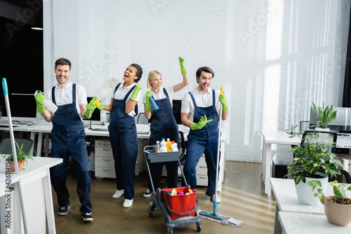 Excited multiethnic cleaners in uniform looking at camera in office