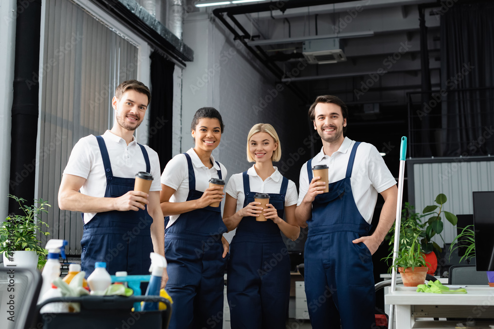 Interracial cleaners with paper cups looking at camera near detergents on blurred foreground in office Interracial cleaners with paper cups looking at camera near detergents on blurred foreground in office