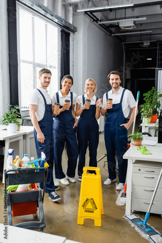 Cheerful multiethnic cleaners holding coffee to go near detergents in office