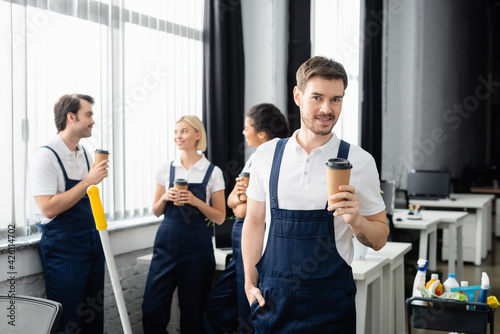 Smiling worker of cleaning company with paper cup looking at camera near multiethnic colleagues in office