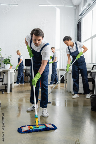 Cleaner in overalls washing floor near multiethnic colleagues in office