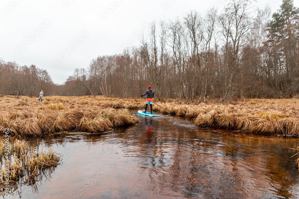 Obraz premium Young men paddle with SUP or stand up paddle board in small river. concept of harmony with the nature. Stand up paddle boarding - awesome active outdoor recreation.
