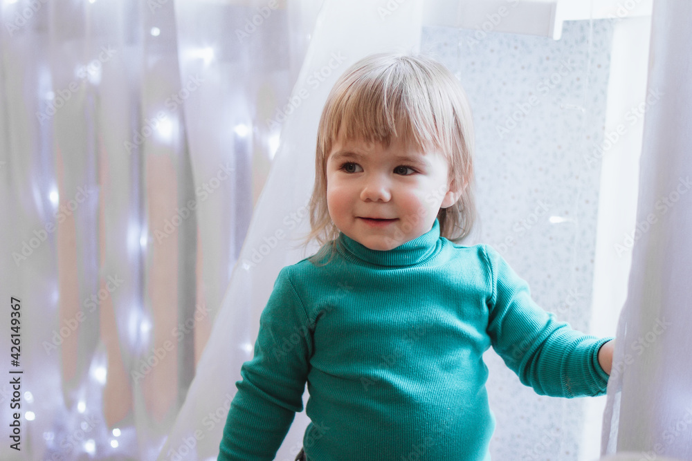 Portrait of smiling little girl with blond hair in turquoise sweater on a background of white curtains on a window with lights. Portrait of smiling little girl with blond hair in turquoise sweater on a background of white curtains on a window with lights.
