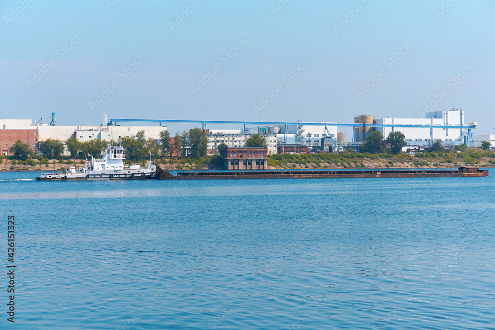 towboat pushes dry bulk cargo barge on the river against the backdrop of an industrial landscape on the shore