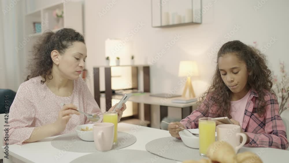 Mother and daughter eating breakfast smartphones in hands, lack of communication Mother and daughter eating breakfast smartphones in hands, lack of communication