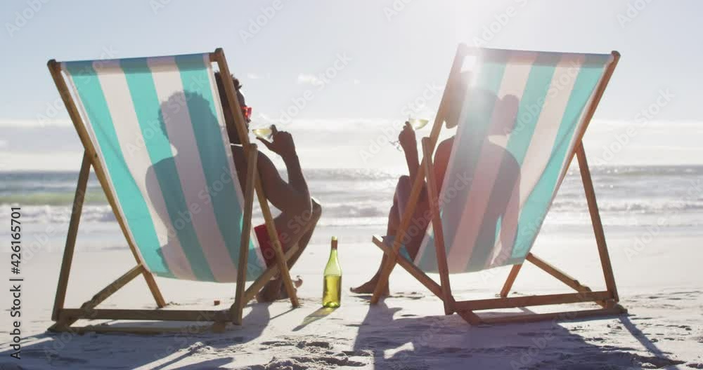 African american couple drinking wine together sitting on deck chairs at the beach