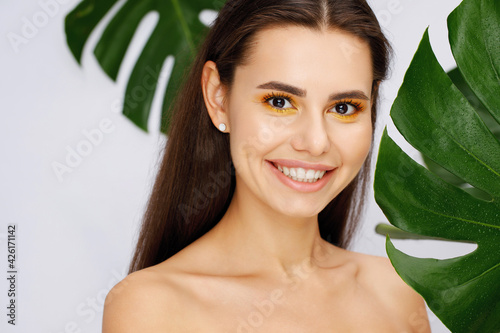 Portrait of young beautiful woman with healthy glow skin holds green tropical leaf and covers part of her face. Model with bright modern yellow makeup. Gray background.