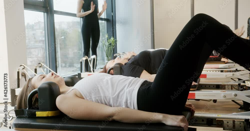 A group of young adult women train in Pilates on a reformer simulator in a bright, modern room by the window. A group of young adult women train in Pilates on a reformer simulator in a bright, modern room by the window.