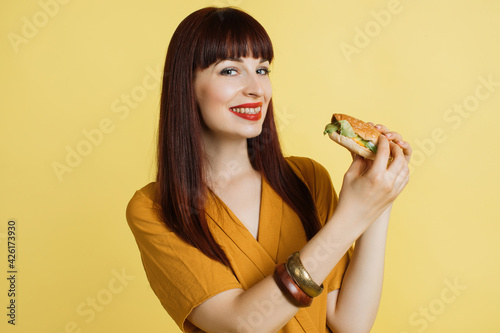 Emotional smiling happy beautiful red haired pretty woman in yellow blouse on yellow studio background, holding fresh tasty cheese burger. Fast food, jank food concept. photo