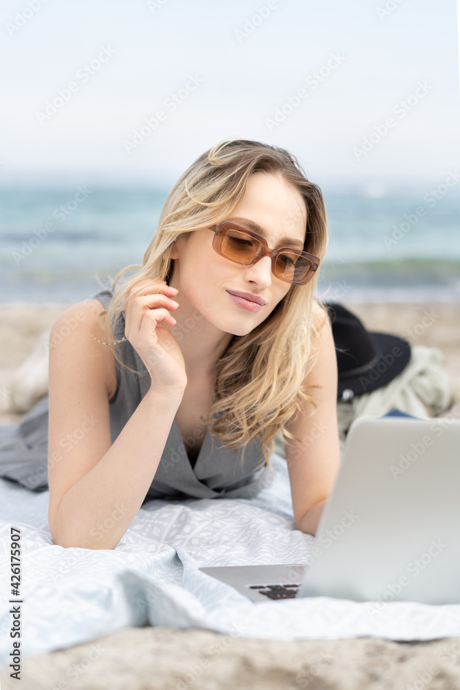 Modern business woman working remotely on the laptop computer wearing sun glasses lying at the beach  