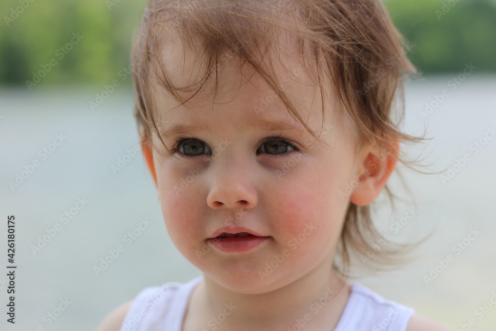 Shining little white girl with gray eyes and curly short hair in a T-shirt stands on the shore of the lake