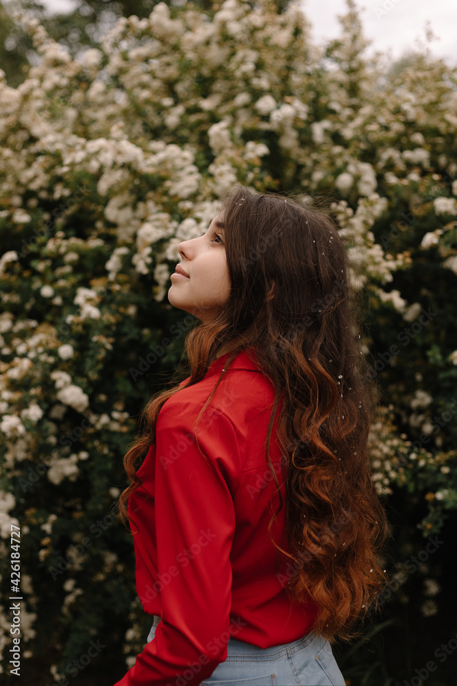 Back view of a beautiful young girl with curly red hair standing near the blooming bush Back view of a beautiful young girl with curly red hair standing near the blooming bush