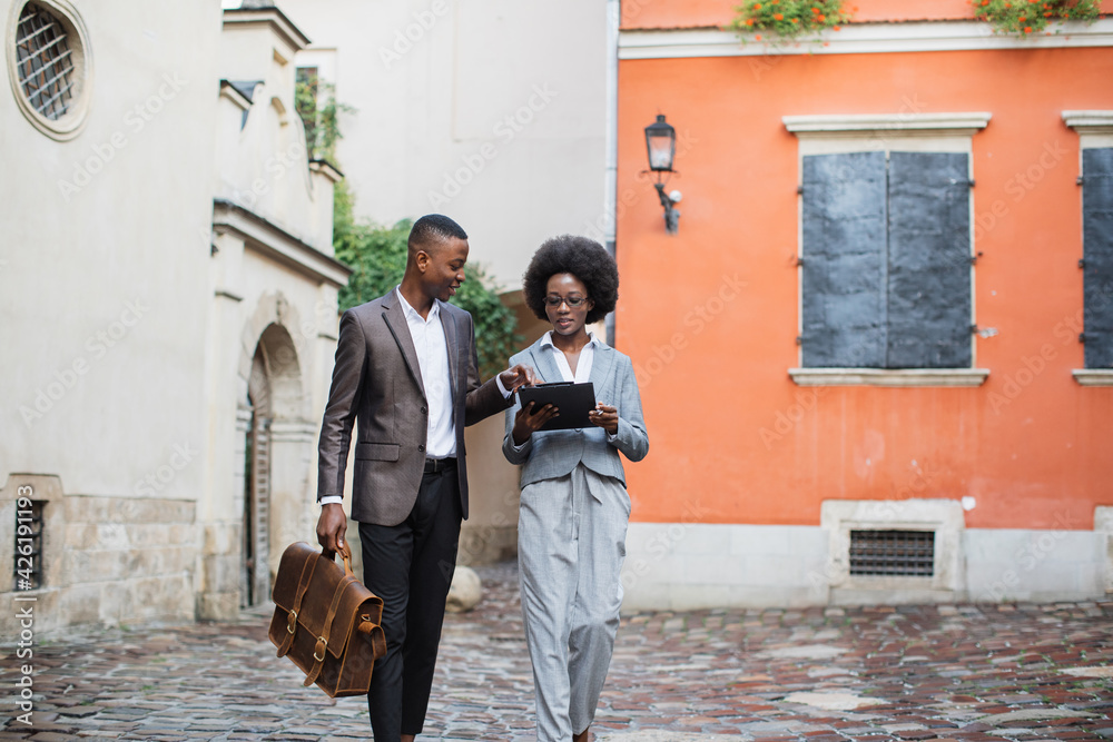 Full length portrait of two african business people in formal clothes walking and talking on city street. Handsome young man carrying brown briefcase, woman holding clipboard.