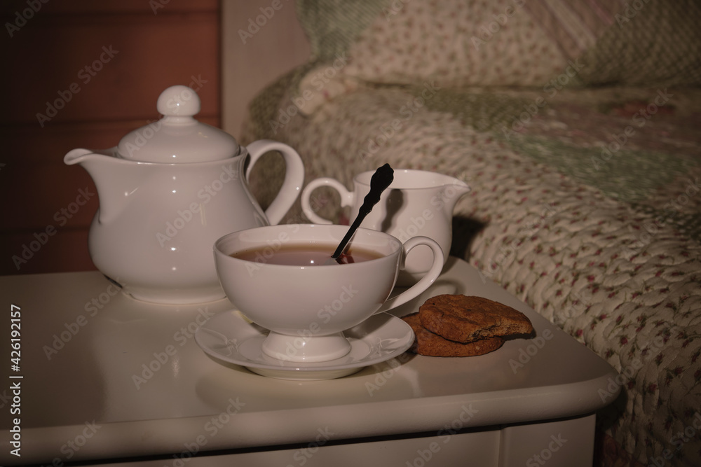A cup of tea, a kettle and a plate of cookies on a small table by the bed in a small cozy bedroom in the morning sunlight. Everything for a good mood. High quality photo