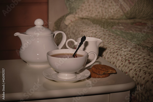 A cup of tea, a kettle and a plate of cookies on a small table by the bed in a small cozy bedroom in the morning sunlight. Everything for a good mood. High quality photo