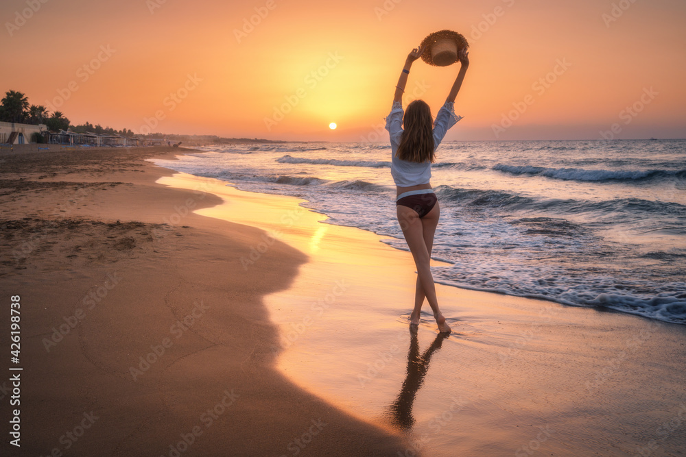 Beautiful young woman with raised up arms with straw hat on sandy beach at colorful sunset. Sea with waves. Summer travel and resort. Happy slim girl in bikini on the tropical sea coast. Lifestyle